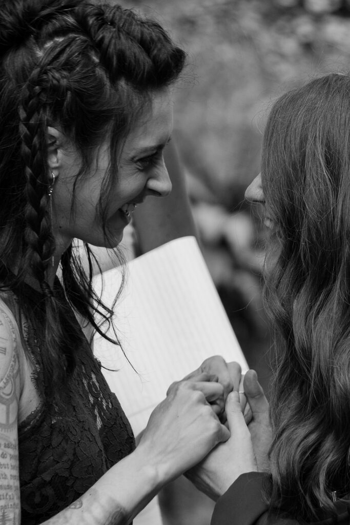 Black and white image of two brides grinning at each other, holding hands during their vow exchange at their elopement.