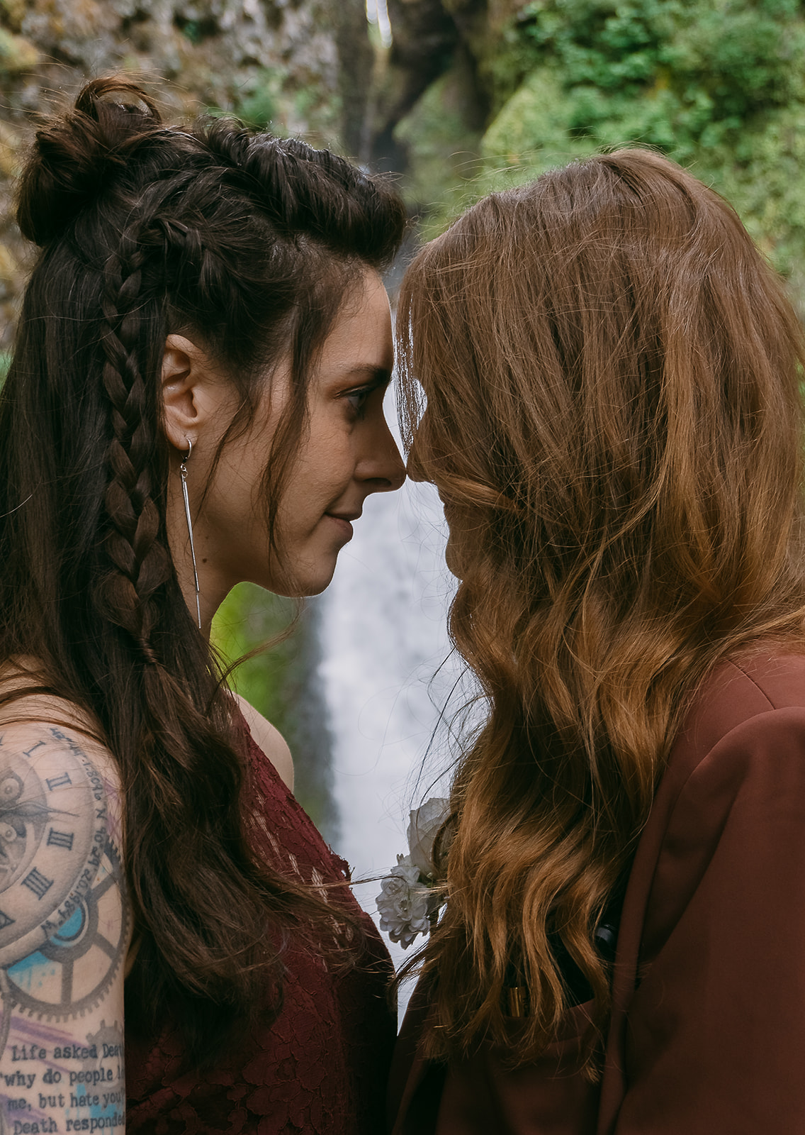 Close up image of two brides looking at each other nose to nose, with the details of their hair featured (one bride has a long braid running down the side of he head, and the other has a long cascade of red curls), in front of a plunging waterfall surrounded by mossy rocks.