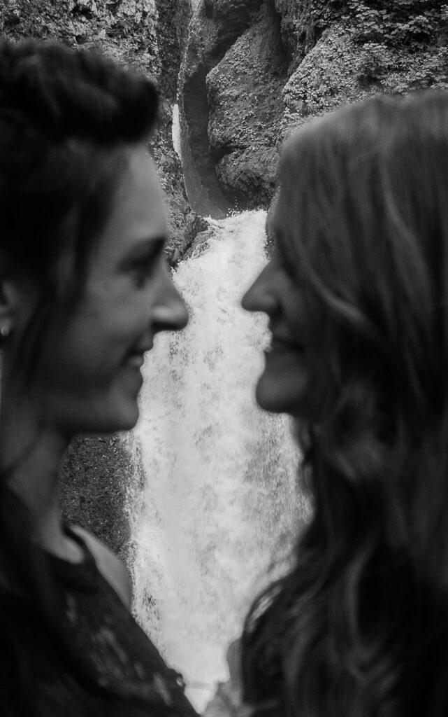 A black and white image of a close up of two brides smiling at each other; their faces are purposefully out of focus, with the waterfall behind them in focus.