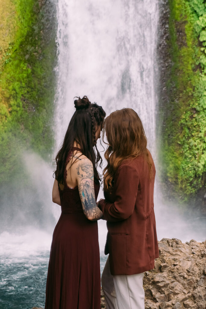 Two brides in maroon and rust colored wedding attire hold hands after their elopement ceremony under a waterfall in Oregon.