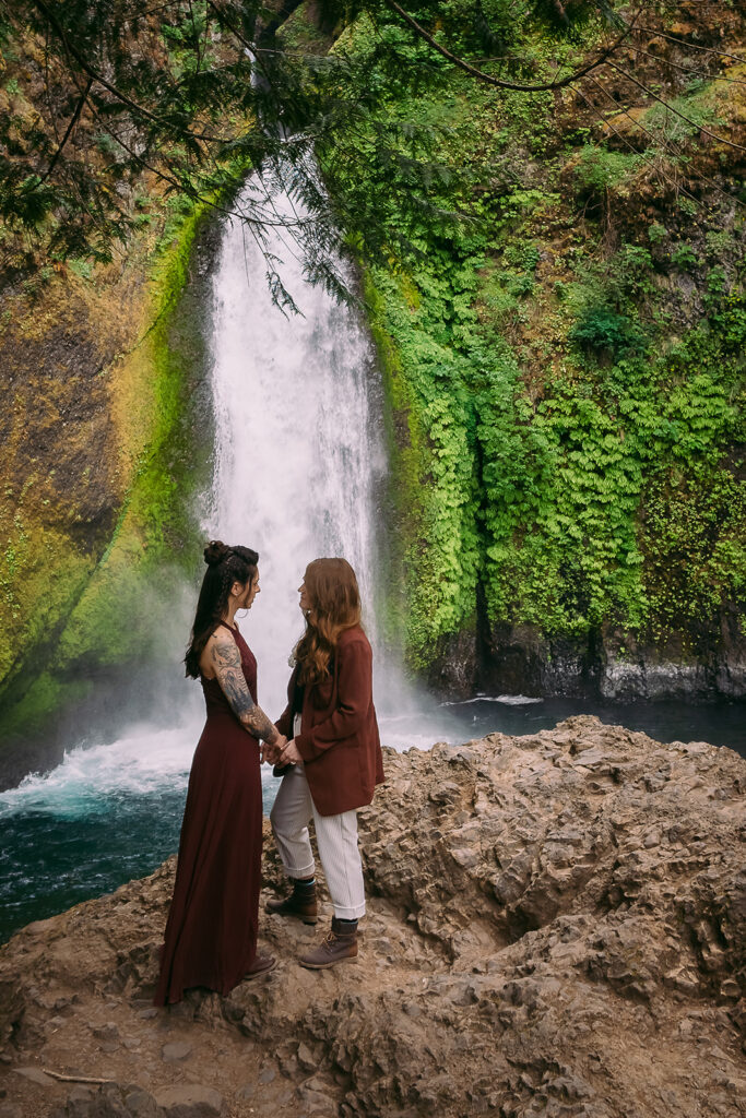 Two brides in untraditional wedding attire hold hands in front of Wahclella Falls in Oregon, after their elopement ceremony.