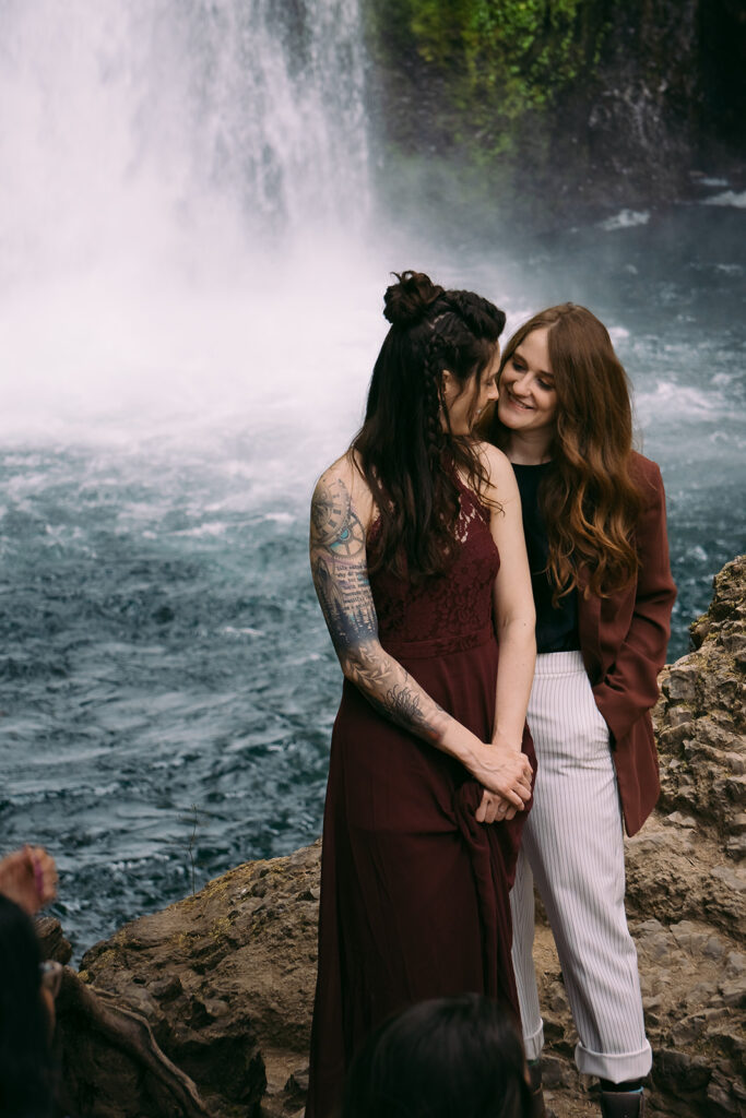 The two brides from the blog post stand side by, looking at each other in front of a blue pool of water where Wahclella Falls falls into.