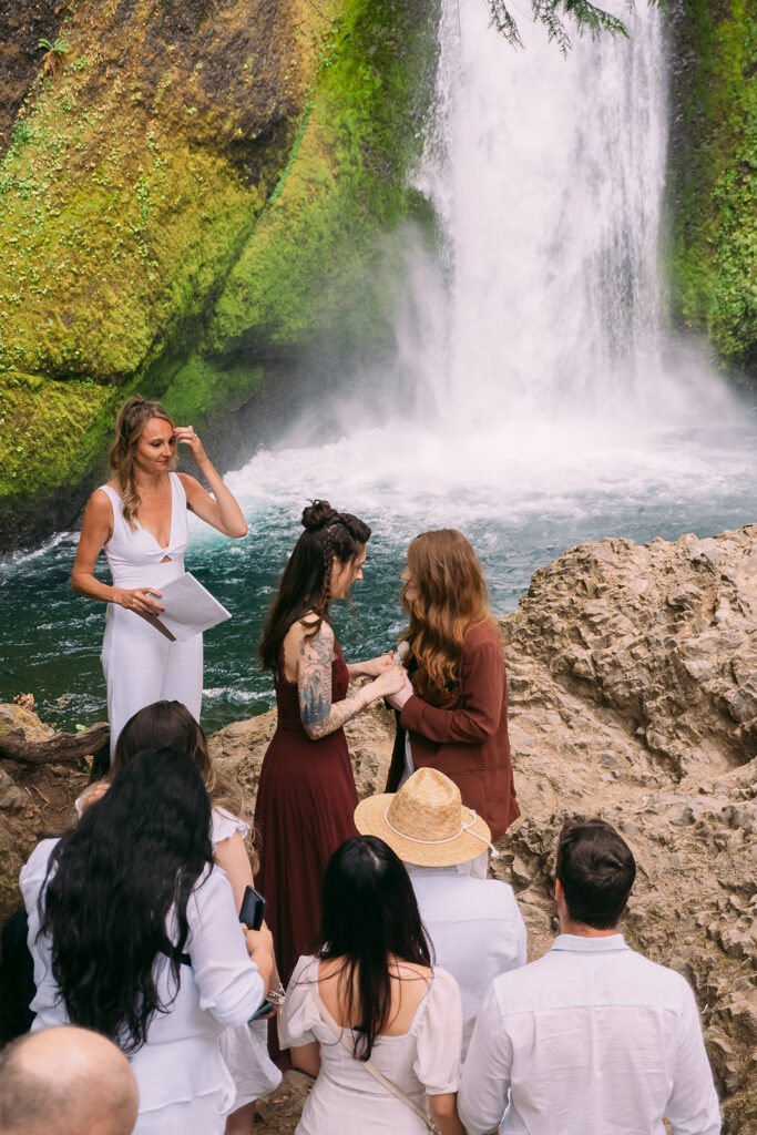 Two brides hold hands in front of a waterfall gushing into a pool of blue water, surrounded by moss covered rocks, as their guests, dressed in white, watch them exchange their vows.