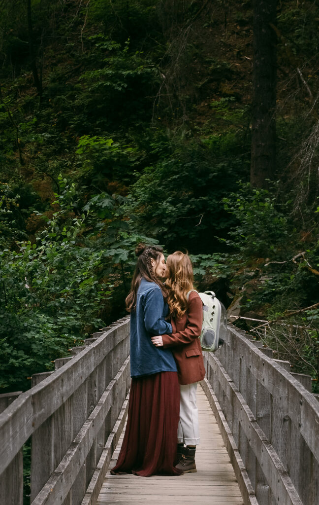 Two brides in reddish wedding clothes kiss on a bridge in the forest.