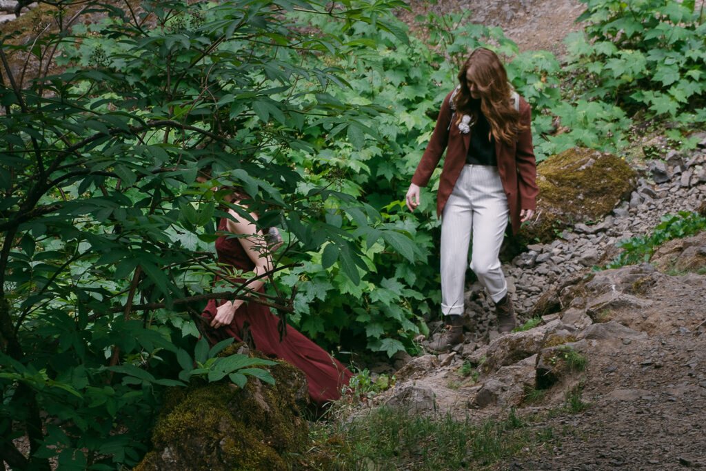 The two brides hike down a rocky slope among the greenery after their elopement ceremony in the forest.