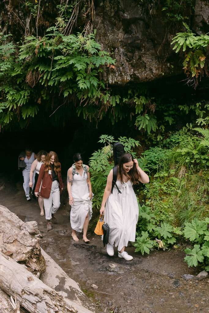 Elopement guests, dressed in white, hike out from under a fern-covered rock  overhang.