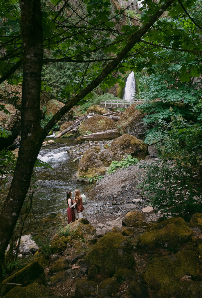 Two brides stand by a river with Wahclella Falls in the background and a big leaf maple tree in the foreground.
